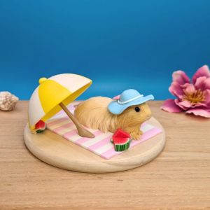 Long-haired cream guinea pig wearing a pastel blue hat with a white ribbon and pink bow, lounging on a stripy white and lavender blanket. A white and yellow umbrella and three slices of watermelon adorn the scene. Guinea pig is set on a wooden base.