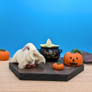 Halloween Guinea Pig dressed as a ghost. There's a cauldron filled with peaflakes and a little cute ghost is sitting on top. There are 3 pumpkins set around them